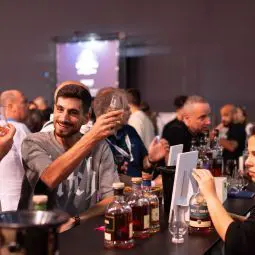 מבקרים טועמים ומדגימים יין וויסקי, לוגו Whisky Live Israel 2025 ברקע חוויית וויסקי בינלאומית בתל אביב.
